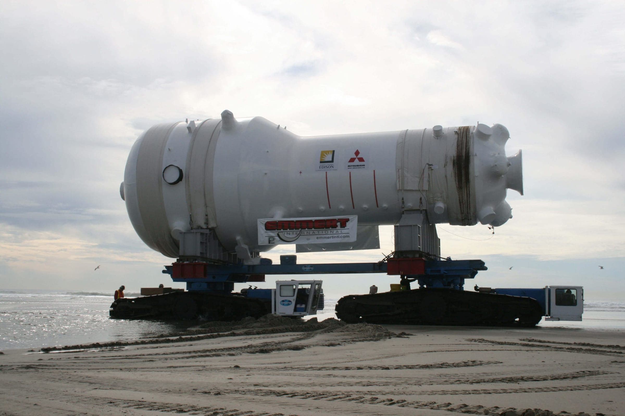 A white replacement steam generator, marked Emmert, carried on a crawler transporter along a wide sandy beach under an overcast sky